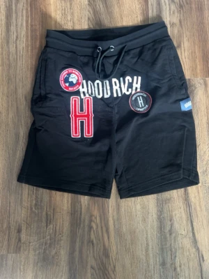 Svarta Hoodrich shorts med patchar - Svarta shorts från Hoodrich med flera patchar och broderade detaljer framtill. Shortsen har snörning i midjan och en relaxed passform. Tillverkade i mjuk bomull, perfekta för sommaren. Stora tryck och loggor ger en streetwear vibe.