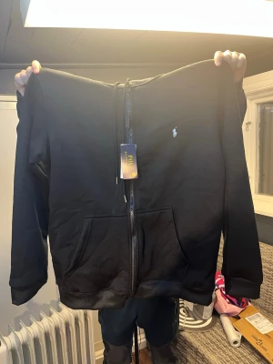 Svart kofta från Polo Ralph Lauren - Säljer en svart hoodie från Polo Ralph Lauren med dragkedja framtill och klassisk logga på bröstet. Hoodien har huva, två stora fickor fram och är tillverkad i mjukt bomullsmaterial. Perfekt för en avslappnad och stilren look.