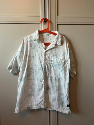 Vit/grön skjorta från Zara - Säljer en luftig skjorta från Zara i vitt med ljusgröna tie-dye detaljer. Skjortan har korta ärmar, klassisk krage, bröstficka och knäppning framtill. Perfekt för en chill och avslappnad stil.