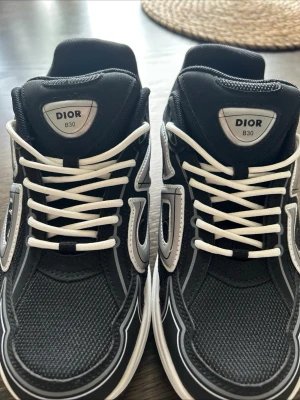 Dior B30 svarta sneakers - Snygga Dior B30 sneakers i svart med vita detaljer och vita snören. Skorna har en sportig design med mesh och läderpaneler samt tydlig Dior-logga på sidan. Tillverkade i Italien och perfekta för dig som gillar exklusiva sneakers.