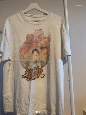 Street Fighter vit t-shirt - Säljer en vit t-shirt med stort Street Fighter-tryck framtill. Motivet visar flera klassiska karaktärer från spelet i färgglada toner. T-shirten har rund hals och korta ärmar, perfekt för dig som gillar retro och gaming. Materialet är mjukt bomullstyg.