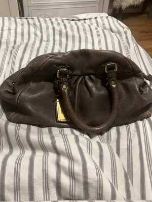 Brun/grön Marc by Marc Jacobs handväska - Intressekoll!! Cool brun handväska från Marc by Marc Jacobs i mjukt läder med guldfärgade detaljer och dubbla handtag. Väskan har ett snyggt mönstrat innerfoder och en metallplatta med logotyp på framsidan. Perfekt för dig som gillar klassisk stil med en twist. Fint skick men finns fläckar på insidan, inget som syns dock. Dessutom lite tecken på användning pga att den är vintage. Detta är den stora väskan.