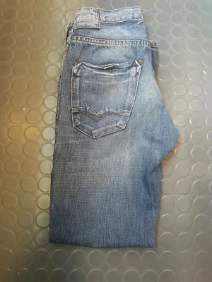Blå raka jeans med slitningar - Säljer ett par blå jeans med raka ben och snygga slitningar framtill. Jeansen har klassiska fem fickor och en cool detalj på bakfickan. Perfekta för en avslappnad och trendig look. Materialet är denim i bomull.