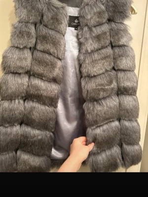 Grå pälsväst från Chelsea - Snygg grå pälsväst från Chelsea i storlek M. Västen har breda, fluffiga paneler i fuskpäls och ett silkeslent foder på insidan. Perfekt för att lyfta din outfit med en lyxig touch och trendig look.