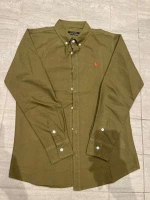 Olivgrön skjorta från Polo Ralph Lauren - Klassisk olivgrön skjorta från Polo Ralph Lauren med orange broderad logga på bröstet. Skjortan har knappar hela vägen, button-down krage och långa ärmar med manschett. Perfekt till jeans eller chinos för en clean look.