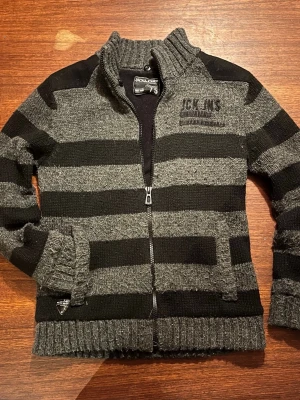 Randig stickad kofta från Jack & Jones - Säljer en stickad kofta från Jack & Jones med breda svarta och grå ränder. Koftan har dragkedja framtill, hög krage och ribbade muddar. Axelpartierna har förstärkningar och det finns fickor på sidorna. Koftan är lite noppig, men det går att fixa. Perfekt nu för våren och för dig som gillar en avslappnad stil! 🌿🍃☀️