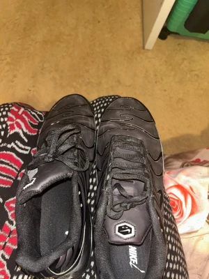 Nike Air Max Plus svarta sneakers - Säljer ett par svarta Nike Air Max Plus sneakers med klassisk TN-logga på hälen och mesh- samt syntetpaneler. Skorna har synlig Air-dämpning i sulan och svarta snören. Snygg design med vågiga detaljer på ovandelen.