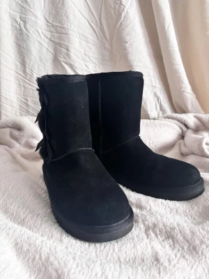 Svarta UGG x Koolaburra boots med rosetter - Mysiga svarta UGG x Koolaburra boots i mocka med fluffigt foder och två dekorativa rosetter på sidan. Klassisk rund tå och mellanhögt skaft. Perfekta för kalla dagar när du vill hålla stilen och värmen.