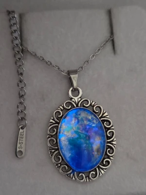 Halsband med blå sten och silverkedja - Unikt halsband med en oval, skimrande blå sten infattad i en detaljerad silverfärgad ram med snirkligt mönster. Kedjan är i rostfritt stål och har en justerbar längd. Efter att ha varit i vanligt dagsljus eller solljus så lyser hänget i ett starkt blått sken i mörker.  Perfekt för dig som gillar färgstarka accessoarer och vill sticka ut. Både kedjan och hänget tål vatten utan att missfärgas . 