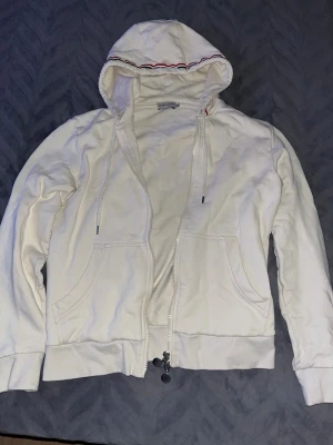 Vit hoodie från Moncler  - Vit hoodie från Moncler med dragkedja och två fickor framtill. Huvan har snören och är dekorerad med en tunn röd och blå rand längs kanten. Mjukt bomullsmaterial och ribbade muddar vid ärmslut och nederkant. Snygga metall-detaljer på dragkedjan.