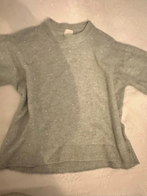 Ljusgrå stickad tröja med pärlor - Mysig ljusgrön stickad tröja med små pärlor insydda över hela plagget. Tröjan har rund halsringning, ribbade muddar och en avslappnad passform. Perfekt för dig som gillar en enkel men ändå lite glittrig look.