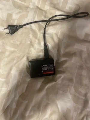 Casio Exilim batteri och laddare - Säljer ett svart Casio Exilim NP-130 litiumjonbatteri (3.7V, 1800mAh) tillsammans med en matchande batteriladdare och strömkabel. Perfekt tillbehör för dig som har en Casio Exilim kamera och behöver extra batteritid eller en extra laddare.