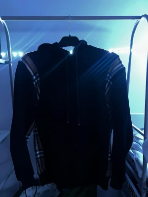 Burberry hoodie  - Snygg svart hoodie från Burberry med klassiska beige, röda och vita rutiga detaljer längs ärmarna. Hoodien har dragsko i huvan och är tillverkad i mjukt material som känns skönt mot huden. Perfekt för dig som vill ha en stilren men ändå unik look.