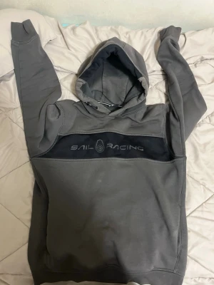 Grå hoodie från Sail Racing - Säljer en grå hoodie från Sail Racing med svart panel och logga broderad över bröstet. Hoodien har huva och långa ärmar, samt ribbade muddar vid ärmslut och nederkant. Tillverkad i mjukt bomullsmaterial för skön känsla.