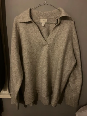 Grå stickad tröja från H&M - Mysig grå stickad tröja från H&M med krage och djup v-ringning. Tröjan har långa ärmar och ribbade muddar vid ärmslut och nederkant. Perfekt för lager på lager och chill stil.