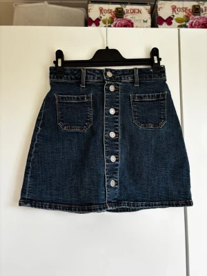 Mörkblå jeanskjol med knappar - Trendig mörkblå jeanskjol med raka snitt, framfickor och knappar längs hela framsidan. Klassisk denimlook som passar perfekt till sneakers eller boots. Kjolen har hög midja och bälteshällor för extra stylingmöjligheter.