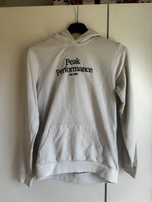Vit hoodie från Peak Performance - Snygg vit hoodie från Peak Performance med svart broderad logga och text 'Est. 1986' på bröstet. Klassisk känguruficka framtill, ribbade muddar och huva. Perfekt för en avslappnad och sportig stil. Materialet känns mjukt och bekvämt.