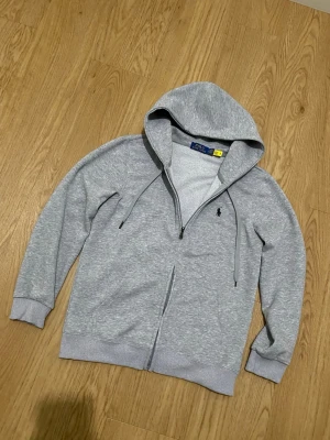 Grå zip hoodie från Polo Ralph Lauren - Snygg gråmelerad hoodie från Polo Ralph Lauren med dragkedja och klassisk huva med snören. Ikoniska svarta polospelaren broderad på bröstet. Tillverkad i mjuk bomullsblandning och har ribbade muddar vid ärmar och nederkant. Perfekt för en chill och stilren look.