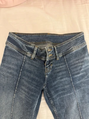 Blå jeans med asymmetrisk midja - Unika blå jeans med tvättad look och asymmetrisk midja som ger en cool twist. Jeansen har raka ben, klassiska fickor bak och fram samt dubbla knappar i midjan. 