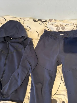 Blå Nike Tech S - Säljer ett blått träningsset från Nike med matchande hoodie och byxor. Hoodien har dragkedja och huva, medan byxorna har dragsko i midjan. Perfekt för gymmet eller chill. Materialet är mjukt och stretchigt för maximal komfort.      Size S