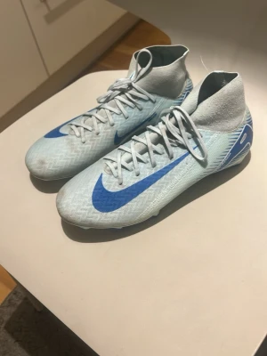 Nike Mercurial blå fotbollsskor - Snygga Nike Mercurial fotbollsskor i ljusblått med blå swoosh och detaljer. Skorna har hög krage för extra stöd runt ankeln och är tillverkade i ett lätt syntetmaterial som ger bra bollkontroll och passform. Perfekta för dig som vill ha stilrena och snabba skor på planen.