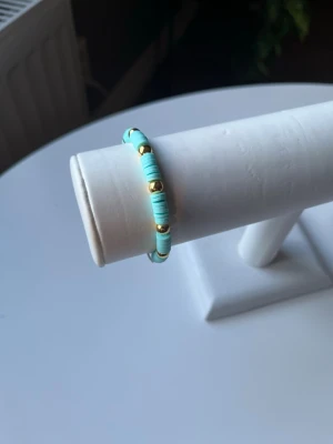  Turkos somrigt armband med lite gulddetaljer  -  Ett turkos armband med lite gul detaljer som har elastiskt band så att det är lätt att ta på av det är även han gjort.