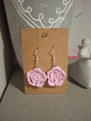 Rosa rosformade örhängen - Säljer ett par handgjorda örhängen i form av rosa rosor. De har krokar i silver och själva rosorna är i lera.. Perfekta för dig som vill ha något gulligt och unikt till din stil.