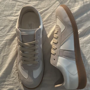 Maison margiela gats vita - Snygga sneakers i vitt och ljusgrått med klassisk retrostil. Ovandelen är i en mix av mocka och slätt läder, med beige skosnören och en brun gummisula som ger en sportig vibe. Perfekta för dig som gillar stilrena och tidlösa sneakers.