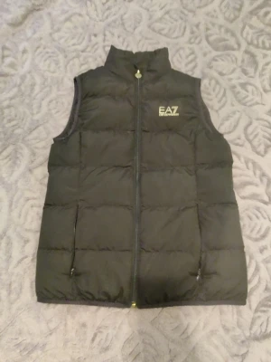 Svart dunväst från EA7 Emporio Armani - Snygg svart dunväst från EA7 Emporio Armani med hög krage och dragkedja framtill. Västen har två fickor med dragkedja och diskret EA7-logga på bröstet. Quiltad design som ger en sportig och modern look. Perfekt för lager-på-lager och streetwear.