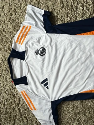 Real Madrid tränings t-shirt Adidas - Snygg vit Real Madrid tränings t-shirt från Adidas med marinblå detaljer och orange ränder på axlarna. Tröjan har klubbens logga på bröstet och klassiska Adidas-märket. Perfekt för fotbollsträning eller sportiga dagar.