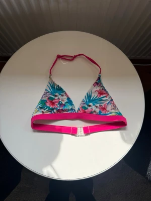 Färgglad trekantsbikinitopp med blommor - Snygg trekantsbikinitopp och underdel med tropiskt blommönster i blått, rosa och grönt. Rosa band och kant, justerbar i nacken och spänne i ryggen. Perfekt för sommaren och strandhäng! I storlek 158/164, från stadium