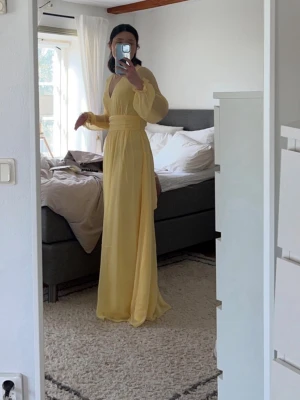 Ljusgul balklänning  - Elegant ljusgul långklänning med v-ringning, markerad midja och lång ärm. Klänningen har en hög slits på sidan och är tillverkad i ett lätt, flytande material som ger en romantisk känsla. Perfekt för sommarens speciella tillfällen.