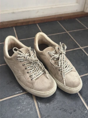 Beige sneakers från Zadie & Volt - Snygga beige sneakers från Zadie & Volt i mocka med extra sulor i och coola vita skosnören med svart text. Diskret stjärndetalj på sidan och färgglad detalj vid hälen. Perfekta för dig som gillar en clean och trendig look.
