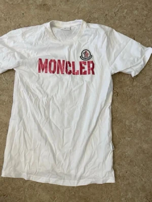 Vit Moncler t-shirt med röd logga - Säljer en vit t-shirt från Moncler med stor röd MONCLER-text och klassisk logga på bröstet. T-shirten har rund halsringning och korta ärmar. Perfekt för dig som gillar stilrena och exklusiva plagg med tydlig branding.