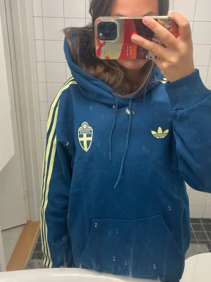 Blå Adidas hoodie med Sverige-märke - Säljer en blå hoodie från Adidas med Sveriges landslagssköld på bröstet och klassiska gula ränder längs ärmarna HELT oanvänd. Perfekt för VM i sommar. Hoddien är unisex men är mer åt M hållet i man storlek. ❗️❗️Pris kan diskuteras ❗️❗️