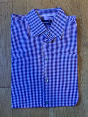 Rutig skjorta från Strellson - Säljer en snygg rutig skjorta från Strellson i shaped fit. Skjortan har ett klassiskt krage, knappar framtill och ett mönster i vinrött och vitt. Perfekt för dig som gillar stilrena detaljer och vill ha en skjorta som sticker ut lite.