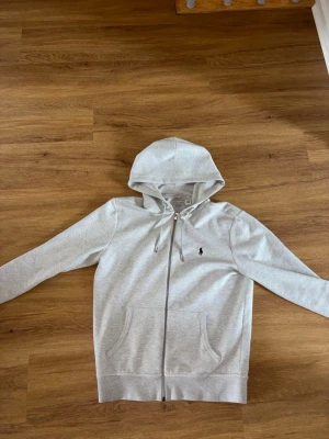Ljusgrå hoodie från Polo Ralph Lauren - Snygg ljusgrå hoodie från Polo Ralph Lauren med dragkedja framtill och klassisk huva med dragsko. Broderad logga på bröstet och två stora fickor framtill. Tillverkad i mjuk bomullsblandning som känns skön mot huden. Perfekt för en avslappnad och stilren look. Nypris ca 1900kr
