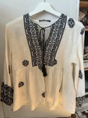 Blus med bredyr - Bohemisk vit blus från Zara med svart broderi längs halsringningen, ärmsluten och utspritt över hela plagget. Blusen har knytband med tofsar vid halsen och är tillverkad i ett lätt, luftigt tyg som ger en avslappnad känsla.