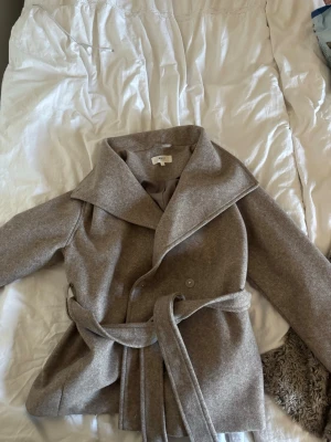 Beige ullkappa med skärp - Stilren beige ullkappa med bred krage och knytbart midjebälte. Kappan har en mjuk, borstad yta och klassisk passform som ger en snygg siluett. Perfekt för kyliga dagar och enkel att matcha med olika outfits.