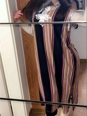 Marockansk traditionell kläning - Färgstark långklänning med vertikala ränder i svart, rosa, gul och vit. Klänningen har snörningsdetalj vid halsen och korta ärmar. Materialet ser ut att vara bomull eller polyester, vilket gör den lätt och bekväm. Perfekt för dig som gillar unika mönster och färg.