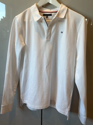 Vit långärmad piké Tommy Hilfiger - Klassisk vit långärmad pikétröja från Tommy Hilfiger med liten broderad logga på bröstet. Skjortan har krage, knäppning vid halsen och är gjord i mjukt bomullstyg. Perfekt för en clean och stilren look.