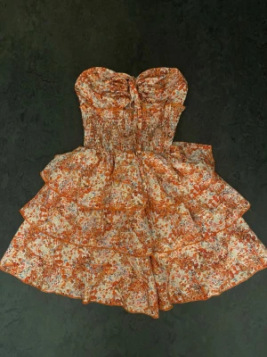 Blommig axelbandslös kortklänning - Supersöt axelbandslös kortklänning med volanger och smock upptill. Klänningen har ett orange och vitt blommönster, knyt framtill och flera lager i kjolen för extra volym. Perfekt för varma dagar och somriga vibes. Klänningen är i storlek Xs. Priset går att diskuteras. 