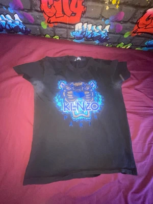 Svart Kenzo t-shirt med tigertryck - Säljer en svart t-shirt från Kenzo med det ikoniska tigerhuvudet broderat i blått och brunt på bröstet. T-shirten har rund hals och korta ärmar. Perfekt för dig som vill ha en statement piece i garderoben.