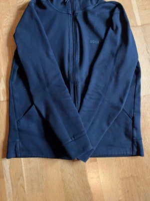 Svart Zip-up med dragkedja från BOSS - Snygg svart hel zip från BOSS med dragkedja framtill och två fickor på sidorna. Diskret BOSS-logga på bröstet. Tillverkad i mjukt material som känns skönt mot huden. Perfekt för en avslappnad och stilren look. Tar emot byten runt om Göteborg!                                                                      Kan sänka priset vid snabb affär