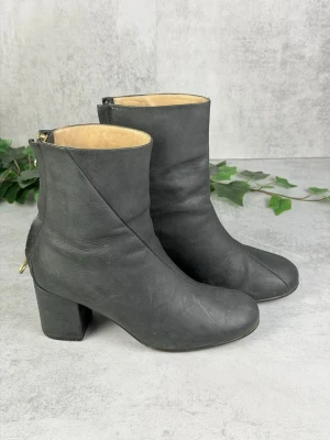 Svarta läderstövletter med klack - Snygga svarta stövletter i äkta läder med rundad tå och kraftig blockklack. Dragkedja baktill i guld för en cool detalj. Insidan är beige och skon har en stilren, minimalistisk design som passar till många outfits.