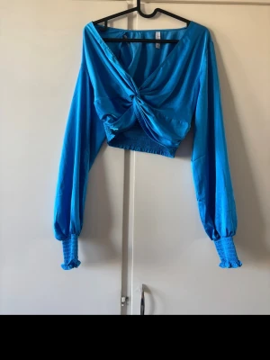 Blå omlottopp med långa ärmar - Snygg blå omlottopp med croppad passform och långa ballongärmar. Toppen har en twistad detalj framtill och breda smockade muddar med volang vid ärmsluten. Perfekt för dig som vill sticka ut med färg och cool design.