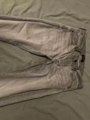 Gråa jeans från Jack & Jones - Säljer ett par gråa jeans från Jack & Jones med klassisk femficksmodell och raka ben. Jeansen har bälteshällor, knappgylf och är tillverkade i ett mjukt denimtyg som ger en avslappnad look.