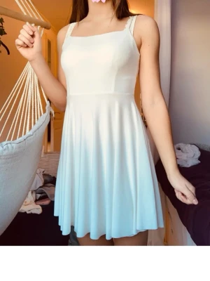 Vit kort klänning med axelband - Säljer en söt vit kort klänning med smala axelband och utställd kjol. Klänningen har en enkel och stilren design med rak urringning och mjukt, följsamt tyg som ger ett fint fall. Perfekt för varma dagar eller sommarens alla tillfällen.