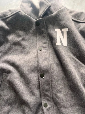 Grå ullkappa från NA-KD - NA-KD Woolblazer / Jacka | Grå | Stl 34 Snygg och mjuk NA-KD-jacka i grå ullig känsla med ett coolt varsity-inspirerat "N"-tryck på bröstet. Svarta tryckknappar ger en modern touch. Perfekt för den som vill kombinera sportig känsla med en mer polerad look. Trendig och mångsidig – passar till allt. 🖤 