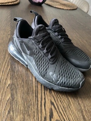 Nike Air Max 270 helsvarta sneakers - Nike Air Max 270 sneakers i helsvart med mesh-ovandel och synlig Air-enhet i hälen. Snygg sportig design med ventilerande material och klassisk Nike-logga på sidan. Perfekta för dig som gillar streetwear och vill ha bekväma skor med modern look.